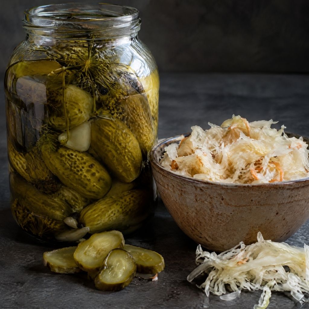 dr-gawlikowska-dieta-przepisy-wielu-z-nas-nie-mieszkajacych-w-polsce-brakuje-naszych-kiszonek-kiszone-ogorki - Dr Gawlikowska - Dr Gawlikowska Glass jar of pickles with garlic and dill next to a bowl of sauerkraut on a dark tabletop.